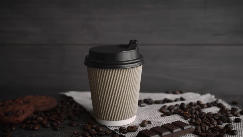 Hand picking up a steaming coffee cup from a table with coffee beans, chocolate, and cookies for a morning caffeine boost