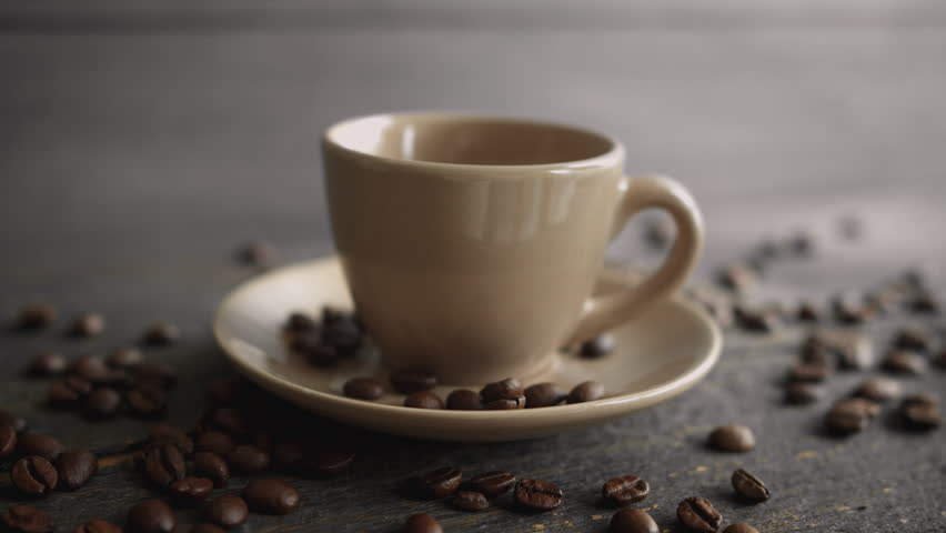 Pouring fresh hot coffee into a cup on a dark table surrounded by roasted coffee beans before a hand picks it up