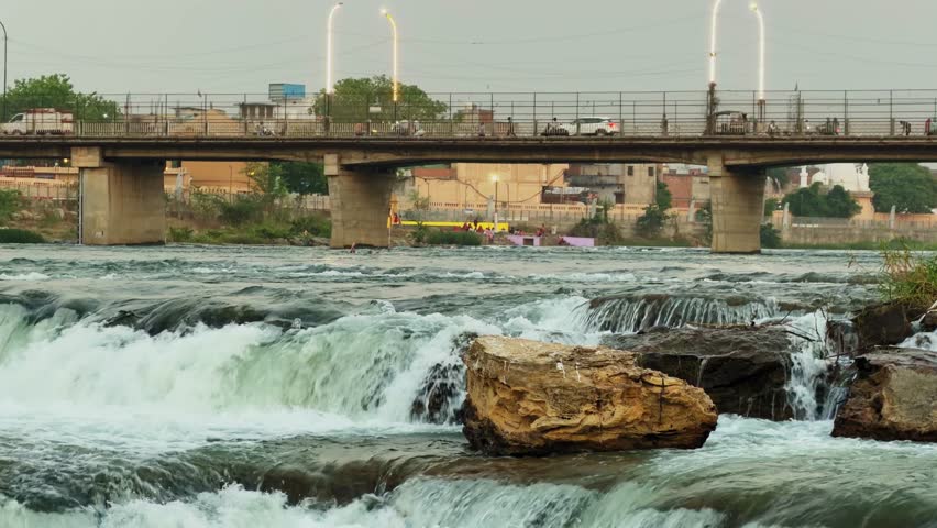 Close view of river rapids cascading over rocks with strong water flow, textured rock formation in foreground and bridge in background under soft natural daylight