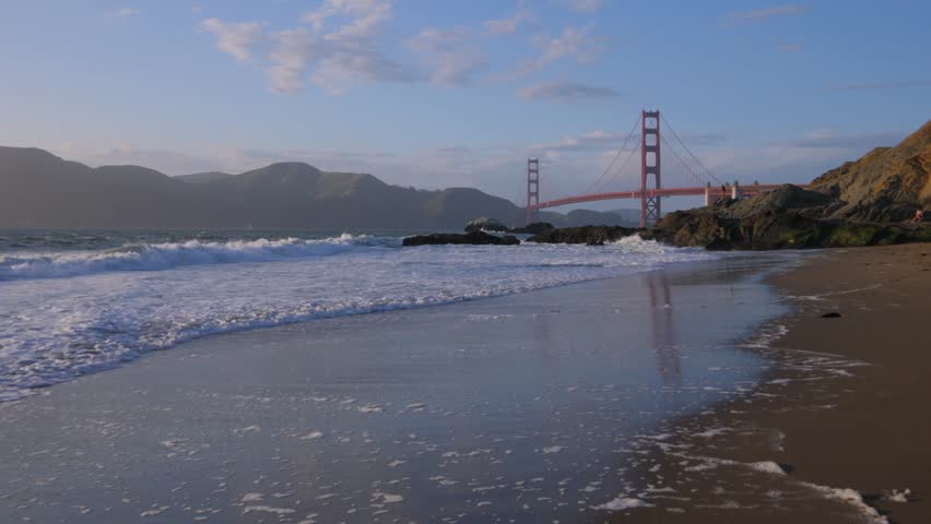This 4K video shows the Golden Gate Bridge as viewed from Baker Beach in San Francisco. The visual documents the suspension bridge spanning the bay entrance with the Marin Headlands topography in the background. The composition illustrates the International Orange structural coating and coastal shoreline, documenting maritime geography and civil engineering for editorial media.