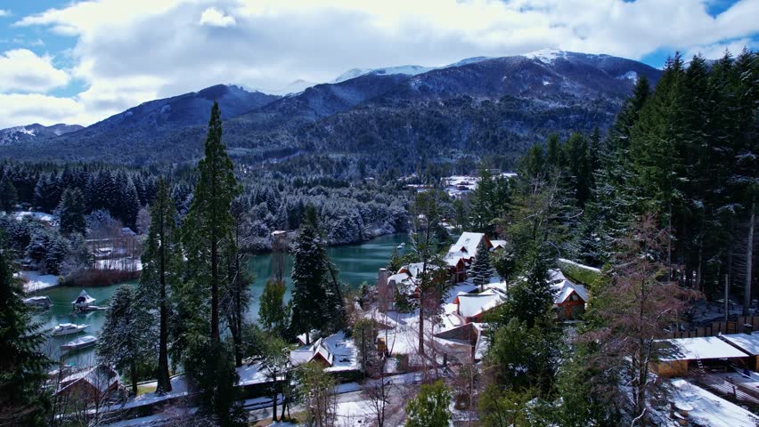 Drone rising vertically while the camera tilts down, transitioning from a panoramic view of the Andes to a high angle shot of a snowy bay with boats on Nahuel Huapi Lake in Patagonia.
