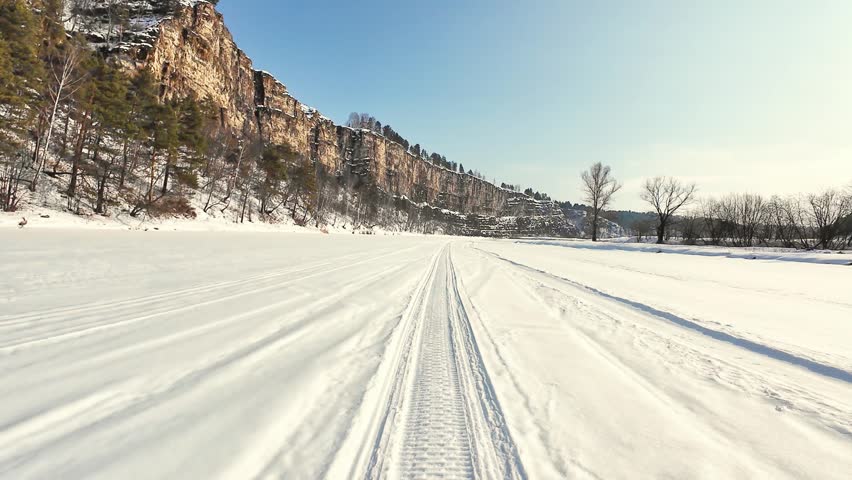 Wide Frozen Track Leading Toward Rocky Cliffs, Parallel Tire Impressions Cut Across Bright Snow, Clear Sky And Distant Bare Trees, Sense Of Open Route And Slow Winter Travel.