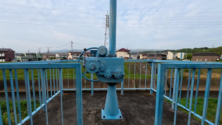 Close-up of a blue mechanical control structure on a platform, with a suburban Japanese landscape in the background, a rural, peaceful and quiet neighborhood 