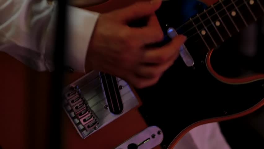 male hands strumming orange electric guitar strings featuring maple neck chrome hardware white shirt sleeve blurred nightclub background atmospheric stage lighting.