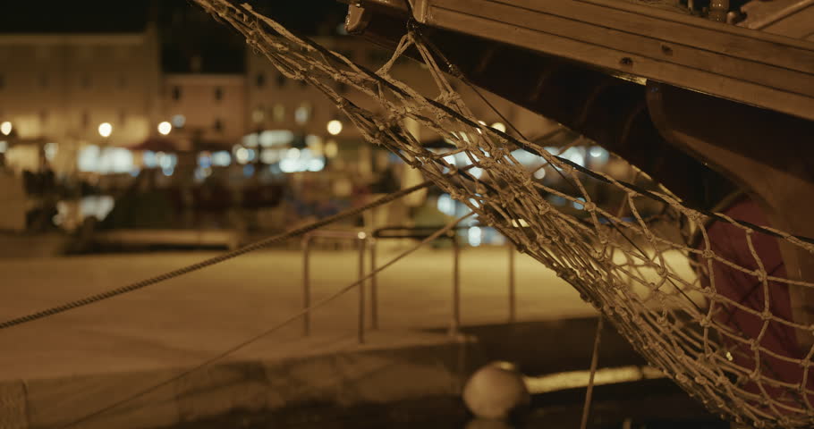 The bow of a wooden boat with rope netting is shown above the water with a waterfront area in the background at night.