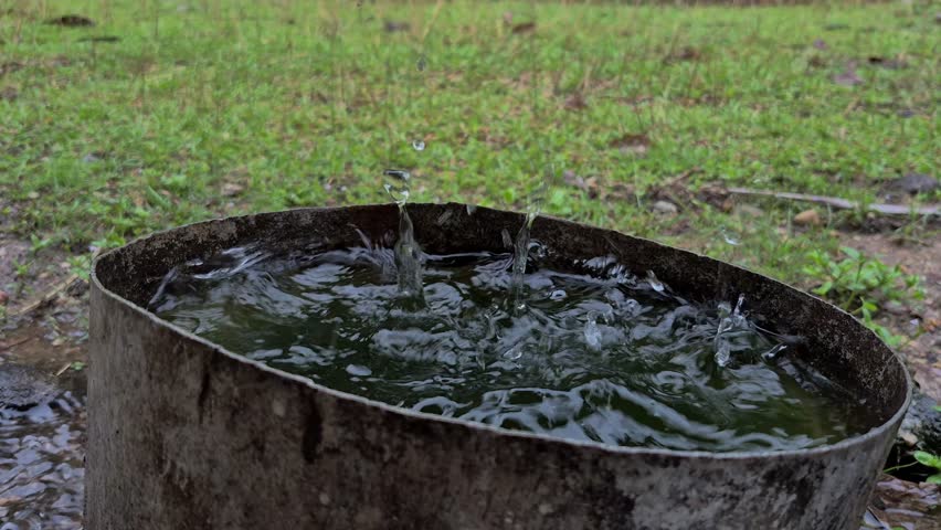 Slow motion raindrops hitting water in a container, creating splashes and ripples in a rural outdoor setting.
