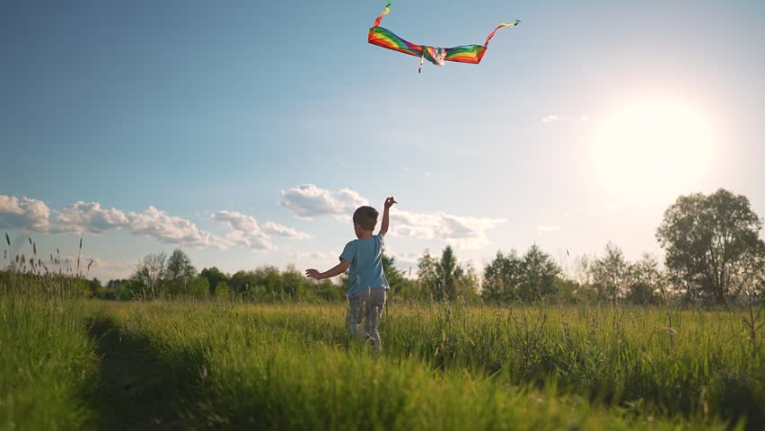 Child running across green field happy. Boy lifts kite flying high. Summer outdoors brings carefree freedom. Kid laughs running wild. Boy enjoys flying kite. Happy child feels outdoors freedom fully.