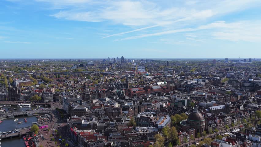 Aerial cityscape showing historic Amsterdam architecture, canals, urban landscape with blue sky on sunny day revealing modern skyline of Netherlands capital in distance