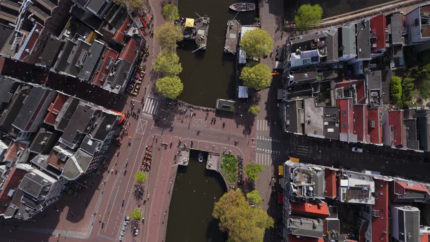 People walking across bridges and streets in Amsterdam, boats mooring in canals and traditional Dutch houses lining waterways, urban life in Netherlands from bird