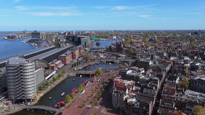 Amsterdam Centraal station and historic city center with canal boats and urban landscape on sunny day, an aerial pan showing famous Netherlands architecture and landmarks