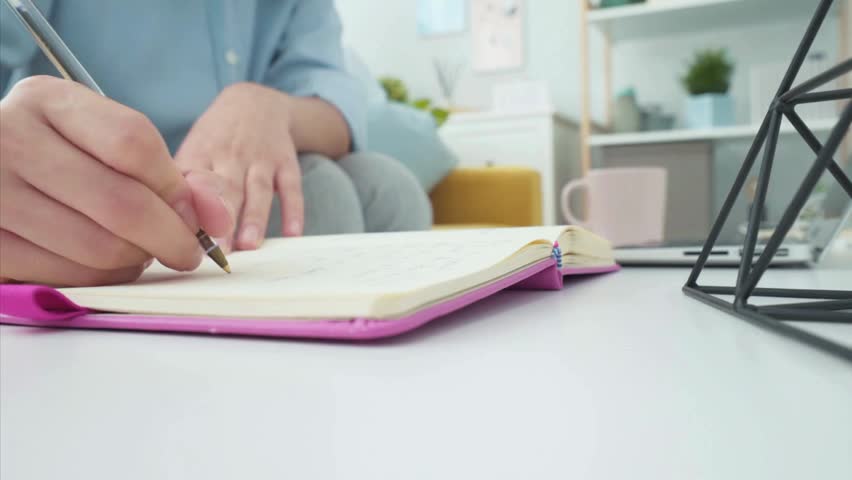 Person writing in a pink notebook on a white desk at home office