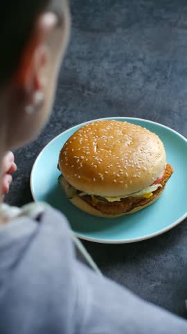 Over the shoulder view of an unrecognizable woman taking a delicious chicken burger from a blue plate, taking a big bite, and putting it back, showing the concept of enjoying fast food. Vertical shot.