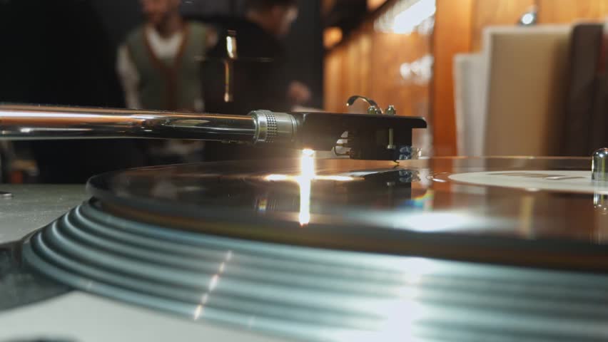 close-up of a vinyl record player with a rotating vinyl record, a tonearm with a pickup needle, DJ hands changing the record, remote control, bar
