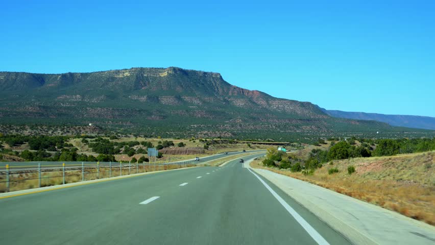 A scenic road trip on a historic highway, likely in the Southwestern United States, possibly Route 66, New Mexico. The photo shows a divided highway running through a dry, elevated landscape with sparse vegetation. In the background, a large mesa or plateau mountain dominates the horizon under a clear blue sky. 