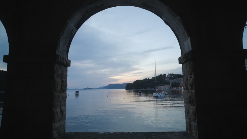 Woman walking beneath a stone archway overlooking Cavtat harbour at dusk in Croatia. Calm water, anchored boats and soft evening light create a peaceful Adriatic travel lifestyle scene.