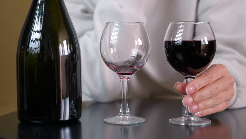 Woman drinks wine alone. Unrecognizable woman holding a glass of red wine, drinking it completely, and placing the empty glass on the black table next to a dark bottle in a static, close-up shot