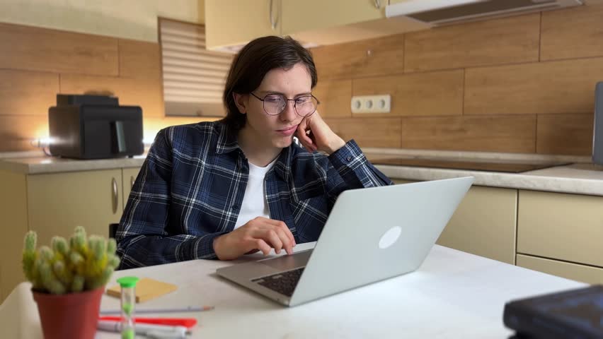 A man sits at a kitchen table, focused on his laptop. The man appears thoughtful as he engages with her laptop in a well-lit kitchen.