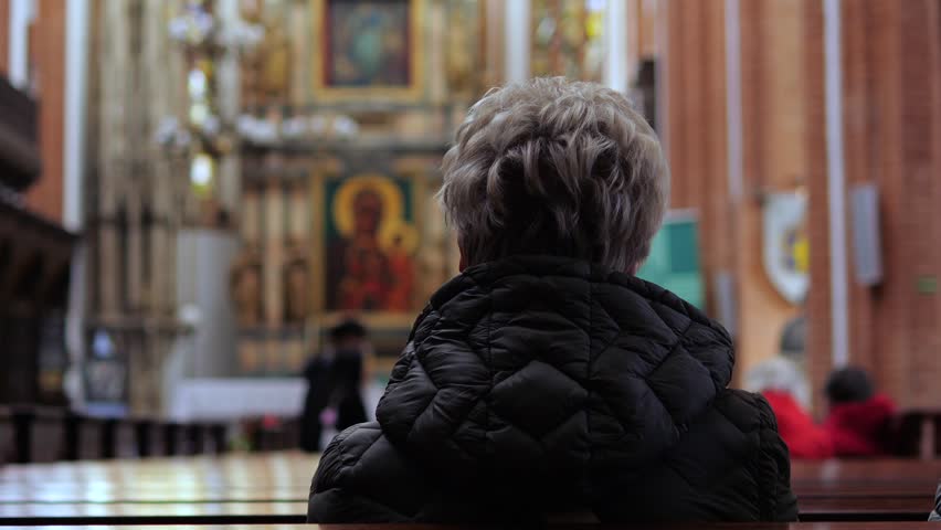 Devout senior woman with gray hair sitting alone on a wooden pew and praying in front of the altar inside a beautiful historic cathedral, showing her deep faith and spirituality