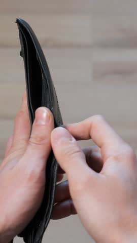 Empty Black Wallet In Hands Showing No Cash, Traveler Inspects Leather Billfold And Card Slot, Closeup On Wooden Floor, Tense Budget Mood, Planning Short Trip, Focus On Lining And Clasp
