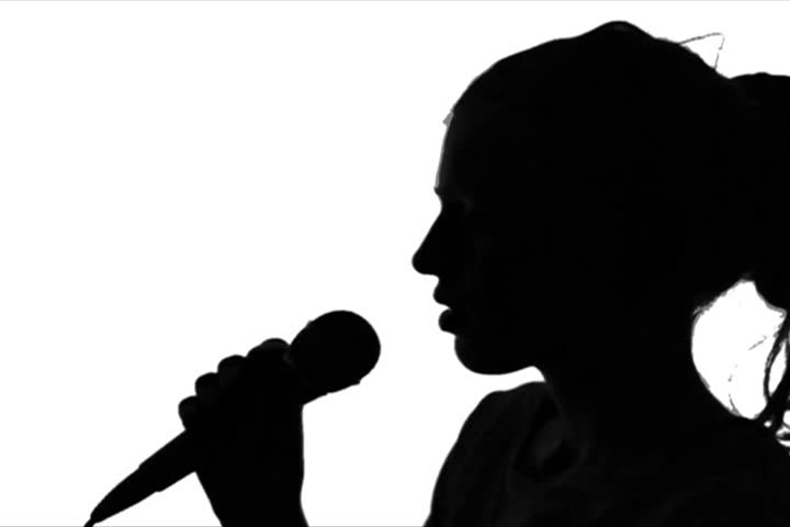Young adult woman holds microphone and sings  against green screen chroma background. Back-lit silhouette. Close-up shot.
