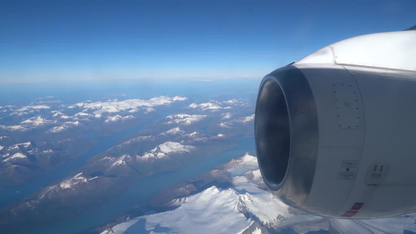 Passenger POV aerial footage flying over the rugged snow-capped peaks and glacial fjords of Patagonia near Bahia Parry, Chile, featuring a commercial jet engine in the foreground