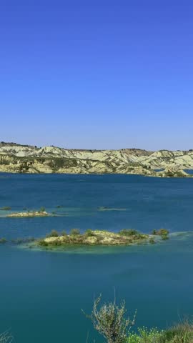 Scenic drone view flying over a tranquil turquoise lake surrounded by arid clay hills and badlands, revealing small green islets under a clear sunny blue sky