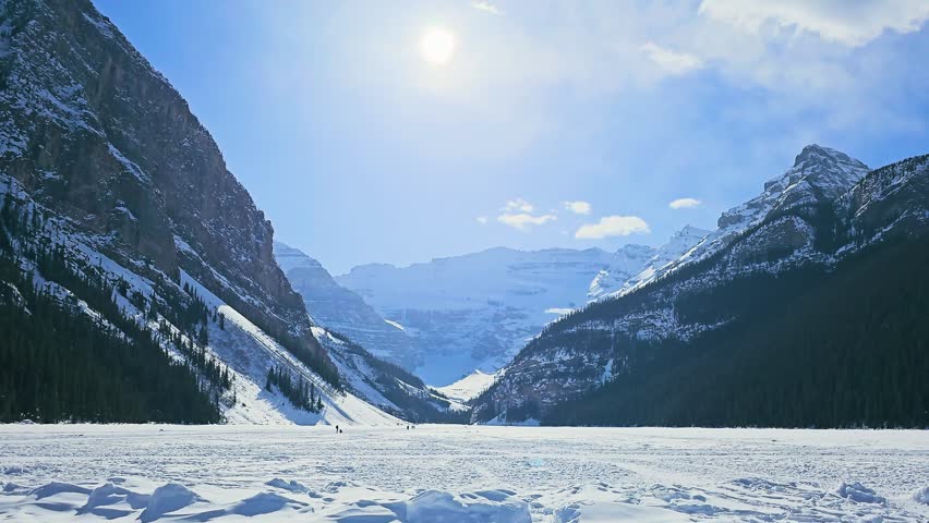 Wide view of Lake Louise in Banff National Park shows snow-covered ground, distant visitors, rugged mountain scenery, dense pines, frozen lake surface, and clear winter light.