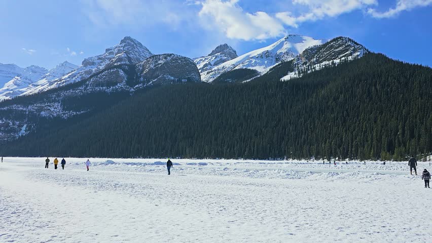 Lake Louise in Banff National Park presents a stunning winter view with snowy ground, forested slopes, towering peaks, distant people, blue sky, and a frozen lake beneath rocky cliffs.