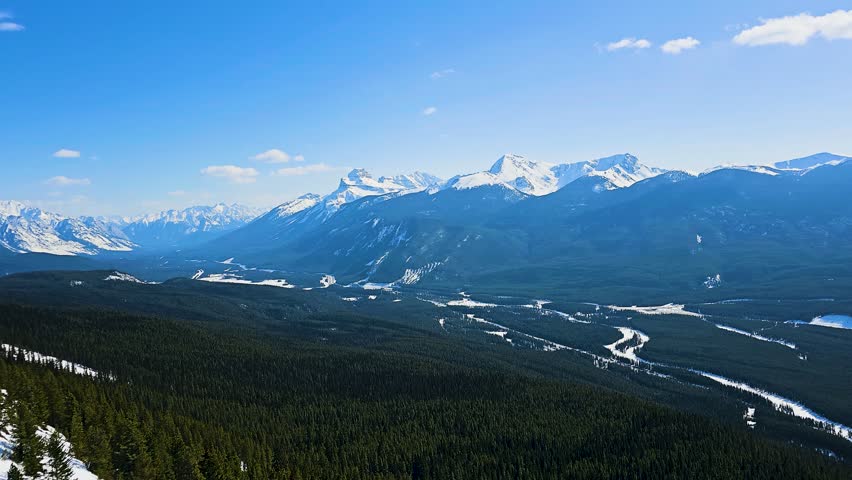 Scenic Banff National Park view with towering snowcapped mountains rising above a vast forest and river valley, showcasing dramatic alpine terrain and classic Canadian wilderness beauty.