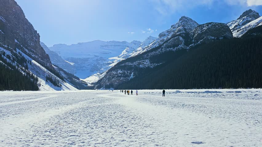 Lake Louise in Banff, Canada stretches across a frozen snowy landscape with towering Rocky Mountain peaks, dark pine forest, distant visitors, bright snow, and crisp blue winter sky.