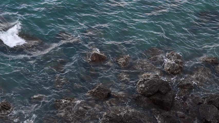 Ocean Waves Crashing on Rocky Shore. Close-up aerial view of dark blue-green ocean waves gently lapping and crashing against a rugged, dark rocky shore. The foamy white water highlights the natural beauty of the coastline.