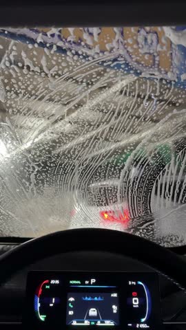 Vertical 4K view from inside a car covered in soap suds, waiting in line for an automatic car wash while workers clean the car ahead.