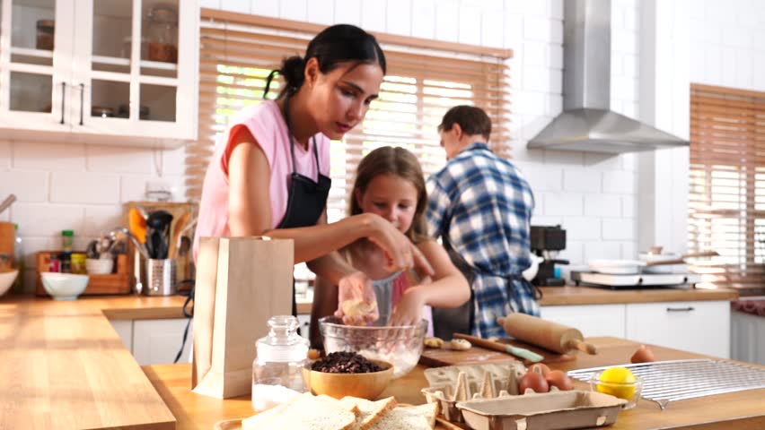 The daughter helps her mother bake cakes and cookies together in the kitchen at home, while the father washes the dishes at the kitchen sink. A family concept.