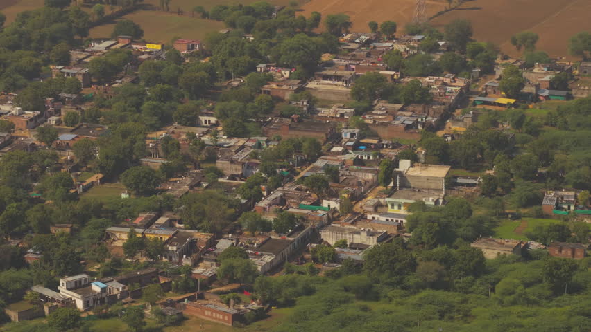 Cinematic aerial shot of a rural Indian settlement with traditional houses, dense green foliage, and surrounding farmland during the day.