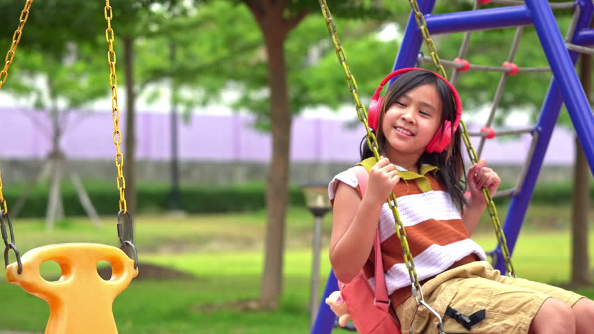 Cute Asian student girl wearing headphones and sitting on a swing while waiting for her mother at school, Back to school and lifestyle concept.