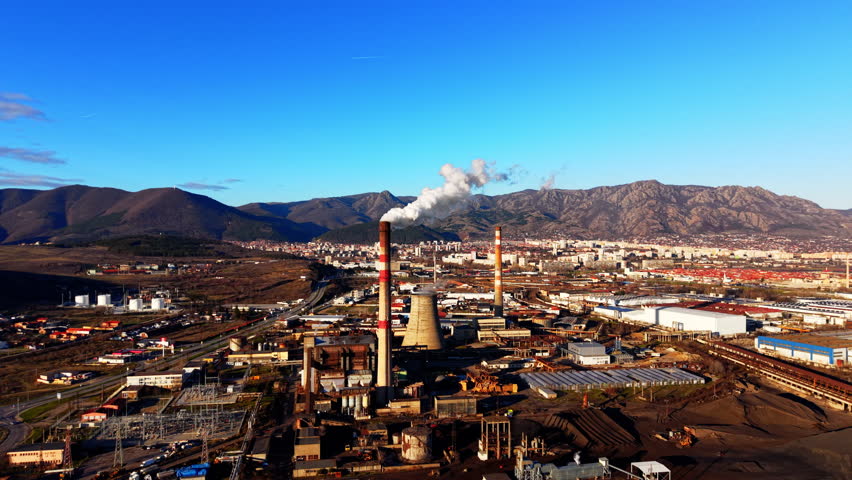 Power station with smoking chimneys, cooling towers, industrial buildings, and urban background under clear blue sky. Aerial view of industrial thermal power plant near city.