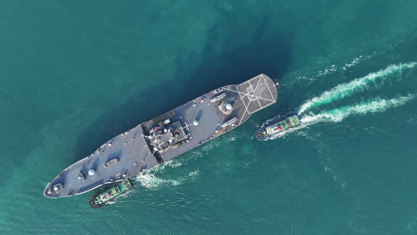 large naval amphibious assault ship being assisted by tugboats in the strategic Strait of Hormuz. Concept of maritime security, naval power projection, and protection of international shipping lanes.