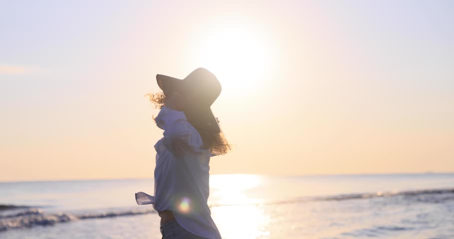 A beautiful woman walking at the beach against the sunset. A happy female enjoying a summer vacation at sea. Freedom and carefree lifestyle concept. Slow motion