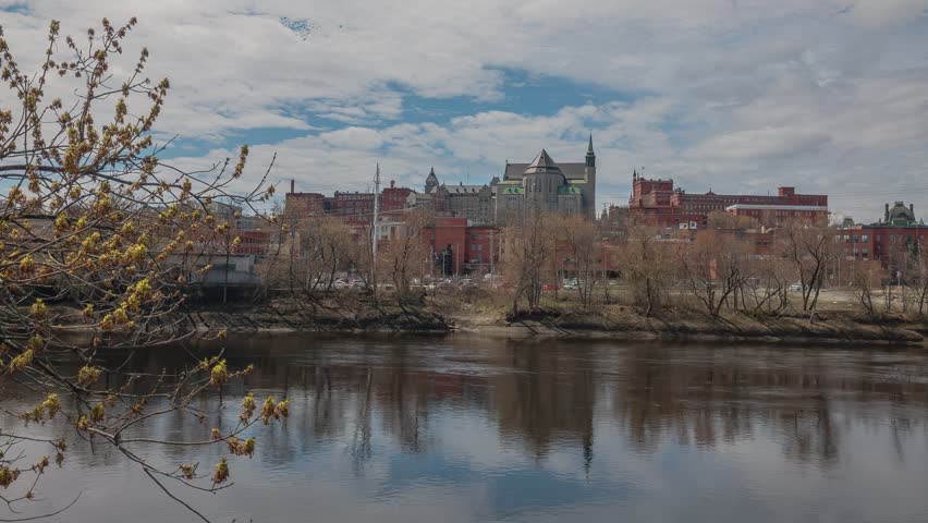Timelapse of downtown Sherbrooke, Quebec, Canada, with historic brick buildings, church spires, and the Magog River in spring. Moving clouds over urban skyline, ideal for travel and city footage.
