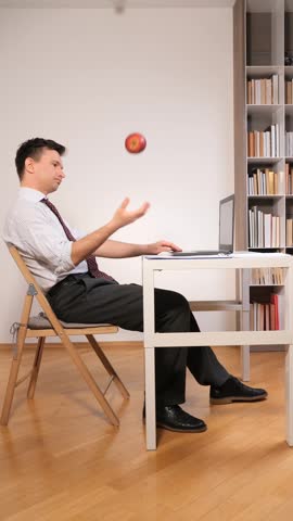 Vertical video businessman sitting at a desk with a laptop, looking bored and tossing a red apple in the air, symbolizing procrastination and a lack of motivation during work