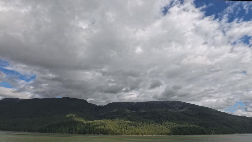Alaska, USA - Mountain range covered in dense forest along the water in Misty Fjords. Cloudy sky with some blue patches above the landscape.