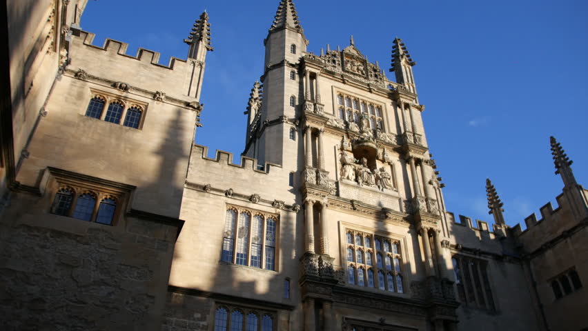 Building facade of Oxford University college with ornate towers, Gothic spires, and sculpted stone figures rising against a clear blue sky in a dramatic low angle view