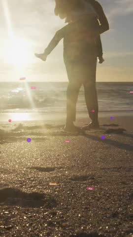 Vertical video: Woman carrying child playing, pivoting from child kick, pink dots on sand marketing. Mom, silhouette, piggyback, backlit, goldenhour, beach, vertical video