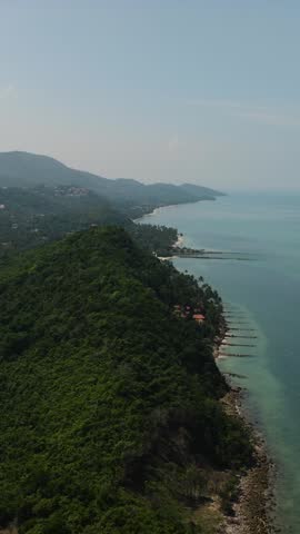 Dense green hills along the coastline meeting the blue ocean waters under a clear sky. Ko Samui, Thailand.