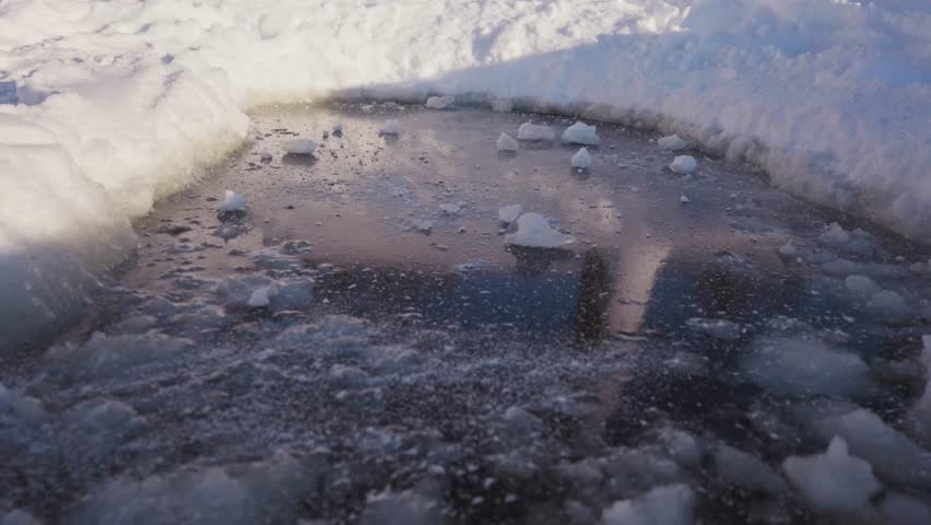Broken chunks of hard snow are scattered across a dark frozen ice patch between white snowbanks, showing rough winter texture, icy ground, and cold outdoor surface details. High quality 4k footage