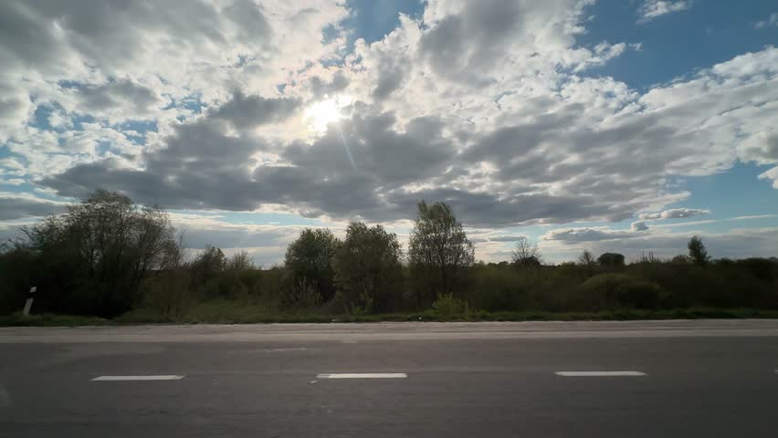 Clouds cover the sky as the sun shines over green fields next to a road in the daylight