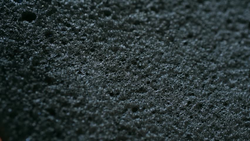 Detailed macro shot of hand spreading white soapy foam onto black porous sponge, with the suds and bubbles slowly absorbing into the surface, illustrating cleaning and hygiene concepts