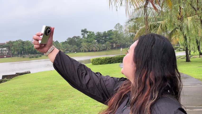 A young Asian woman in a black jacket takes a picture with her digital camera in a park on a rainy day, surrounded by trees and a lake.