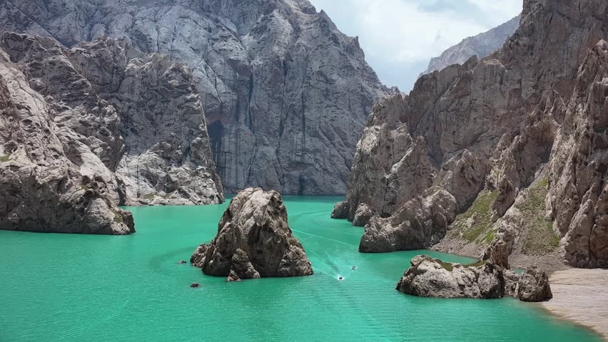 Close-up aerial footage revealing the dramatic texture of jagged rock spires rising from the brilliant turquoise waters of Kel-Suu Lake. The proximity to the rock formations shows the raw geological detail of these ancient canyon walls. Perfect for geology, adventure, and extreme nature documentary content.