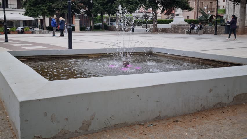 A small decorative fountain sits at the center of a paved public square, gently spraying water upward with soft pink lighting visible beneath the surface